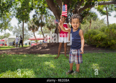 Frère et soeur hispaniques jouer au parc communautaire à l'ombre d'un gros arbre pendant qu'ils détiennent le drapeau américain avec fierté. Banque D'Images