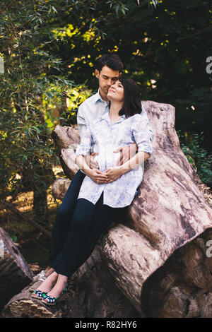 Beautiful pregnant couple en extérieur dans la forêt, tout en étant assis, accolades et embrassades sur une tige de l'arbre avec des feuilles vertes dans l'arrière-plan. Banque D'Images