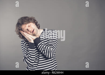Jeune homme en noir et blanc à rayures pour homme veut dormir. Mettre sa tête dans ses mains et ferma les yeux tout en baillant. Debout sur le fond gris. Banque D'Images