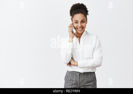 Bonjour, comment allez-vous. Charmant Portrait de femme à la peau sombre confiant in suit holding smartphone, près de l'oreille et le regard avec l'auto-assurance sourire à huis clos, ce mur gris plus de nomination Banque D'Images