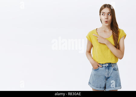 Wow l'air cool. Amusé et excité en femme caucasienne attrayant t-shirt jaune de stupéfaction et lèvres pliantes regardant surprise et pointant vers la gauche avec l'oeil curieux et intéressé sur mur blanc Banque D'Images