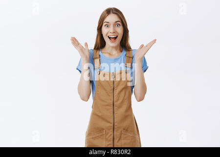 Portrait d'excité et surpris femelle optimiste en salopette marron mignon frappant des mains de stupéfaction et un large sourire heureux sentiment amusé à regarder magnifique perfomance sur mur gris Banque D'Images