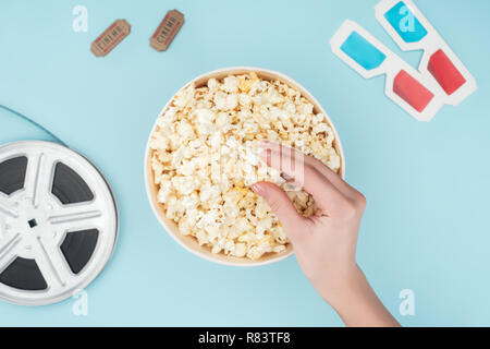 Vue de dessus de bobine de film, lunettes 3D, billets de cinéma et en tenant la main de popcorn sur bleu isolé du godet Banque D'Images