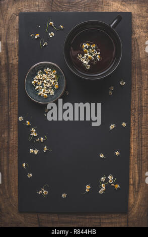 Arrière-plan de thé de camomille sombre avec une tasse de thé, de fleurs de camomille séchées et du miel dans un bol, vue d'en haut. Cadre. Remède pour traiter une vaste gamme de services de santé issu Banque D'Images