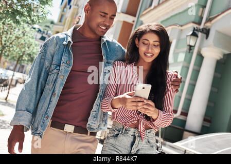 Relation amoureuse. Diverses jeune couple walking on the city street hugging woman browsing internet sur smartphone smiling smiling Banque D'Images