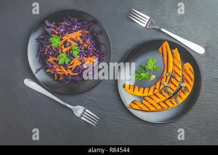 Citrouille en tranches grillées avec le persil et les graines de citrouille, salade de chou violet, les carottes et les graines dans les plaques en céramique noire sur la surface de la pierre noire, haut Banque D'Images