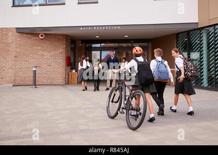 Groupe d'élèves du secondaire en uniforme arrivant à l'école la marche à pied ou vélos d'être accueillis par l'enseignant Banque D'Images