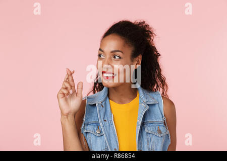 Portrait of a smiling african woman dressed in denim vest isolés sur fond rose, tenant les doigts croisés pour la bonne chance Banque D'Images