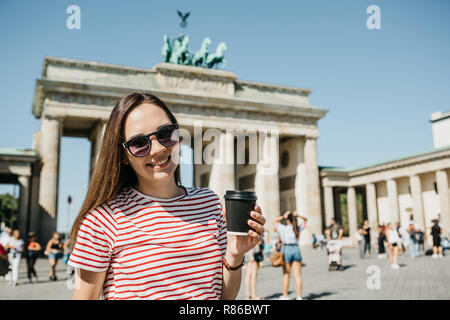 Une fille est titulaire d'un gobelet jetable avec du café ou une autre boisson lors de la marche ou de visites à Berlin, Allemagne. L'avant est la porte de Brandebourg et de personnes non reconnu floue. Banque D'Images