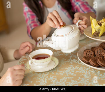 L'on verse du thé noir théière en céramique blanche en Coupe blanche et délicate. Mains de fille Prendre soin de mère en versant un plateau en verre blanc. Banque D'Images