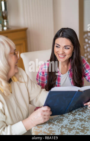 Shot verticale de Cheerful Girl Smiling à sa mère âgée qui lit un livre Hard-Covered Dans Le Salon à la table. Banque D'Images