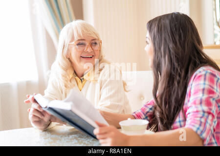 Heureux mère âgée semble heureusement sur sa plus jeune fille qui est titulaire d'un livre ouvert et d'un plateau/bouilloire. Séance de soins. Banque D'Images