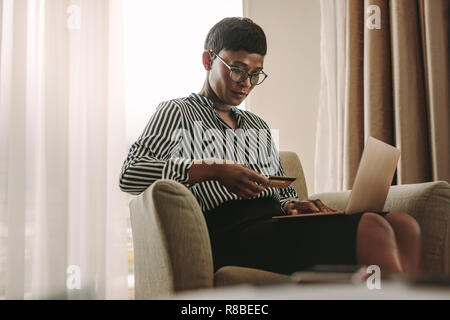 Femme assise sur un fauteuil chambre d'hôtel avec ordinateur portable et carte de crédit. African businesswoman payer sa facture par carte de crédit et un coffre dans chambre d'hôtel. Banque D'Images