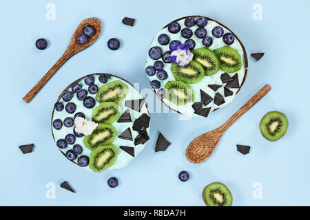 La spiruline bleu de bol, bleuets frais, kiwi et de morceaux de chocolat avec des cuillères en bois servi dans des bols de coco sur fond bleu Banque D'Images