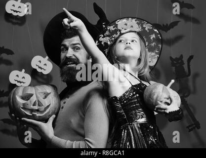 Halloween Party et maison de concept. Père et fille en costumes. Girl pointing up et barbu avec visage curieux posent sur fond gris vert. L'assistant et petite sorcière en chapeaux noirs tenir pumpkin Banque D'Images