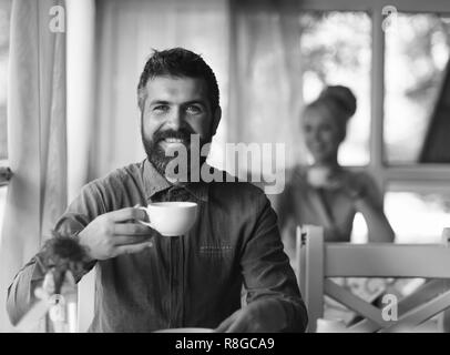 Café du matin temps concept. Homme avec barbe et heureux face détient tasse de boisson chaude sur fond bleu clair cafe. Homme romantique et jolie fille sur derrière ayant une pause. Guy boit du café ou du thé à table Banque D'Images