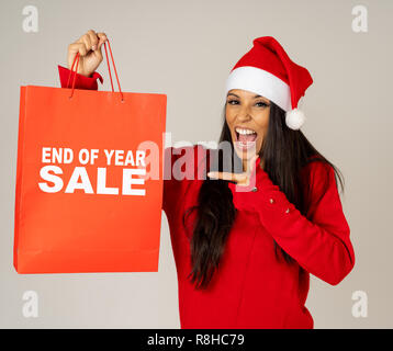 Jeune femme en rouge et santa hat excité avec vente de sacs en papier à fond gris isolé prêt pour les achats de Noël et nouvelle ans consommation ventes Banque D'Images