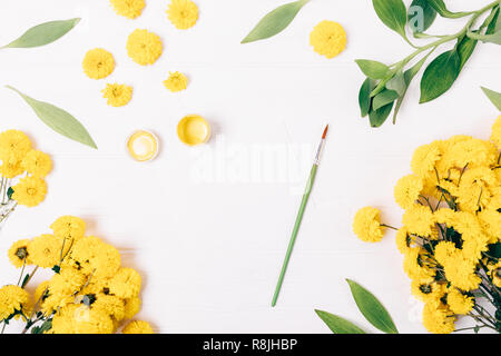 Télévision printemps pondent composition de cartes de vœux vide à côté de la peinture et le pinceau entre les fleurs jaunes et feuilles vertes, vue du dessus sur fond blanc. Banque D'Images