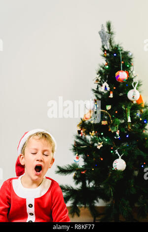 Blonde et beau garçon de 5 ans habillé en père Noël, ennuyé par l'arbre de Noël en attendant l'arrivée du vrai père, fond blanc Banque D'Images