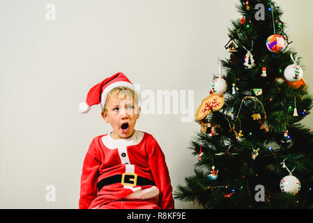 Blonde et beau garçon de 5 ans habillé en père Noël, ennuyé par l'arbre de Noël en attendant l'arrivée du vrai père, fond blanc Banque D'Images