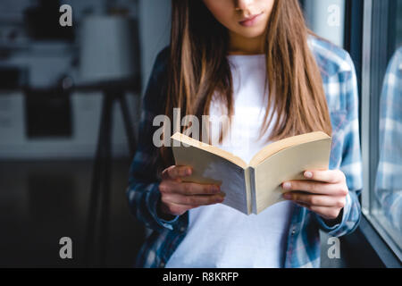 Cropped shot of young woman standing près de fenêtre et lecture du livre à la maison Banque D'Images