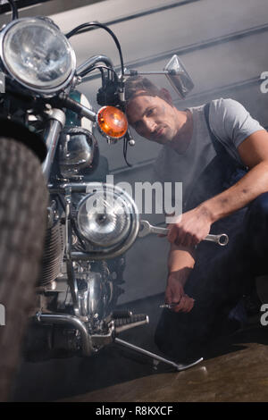 Handsome mechanic holding clé et moteur de moto fixation en garage Banque D'Images