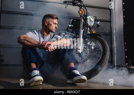 Beau, mécanicien et clé de maintien assis près de moto dans garage Banque D'Images