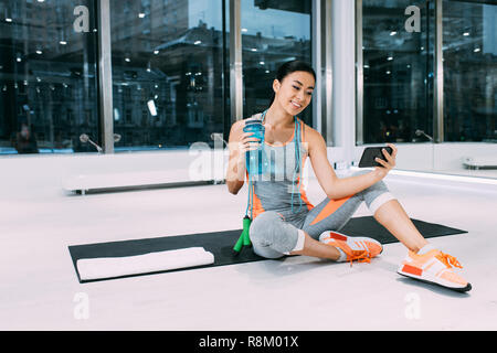 Smiling asian sportswoman assis sur le tapis de fitness sports holding, bouteille d'eau et en tenant à gym selfies Banque D'Images