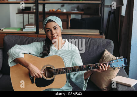 Belle mixed race girl dans chandail turquoise bandeau et playing acoustic guitar on sofa at home and looking at camera Banque D'Images