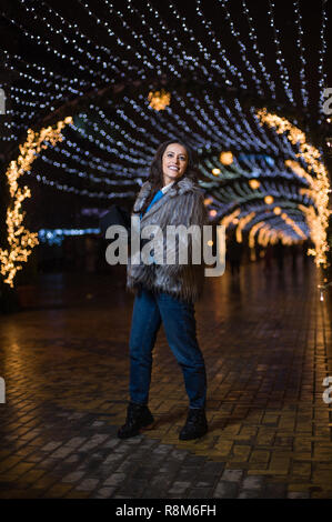 Jolie fille aux cheveux noirs portant un manteau de fourrure, jeans, haut bleu et un chapeau noir, sourire, posant avec des flocons de lumières de Noël à l'extérieur nuit ti Banque D'Images