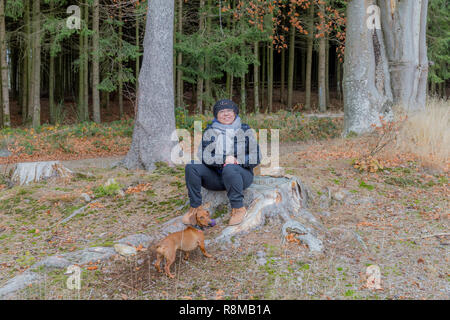 Belle image d'une femme assise sur une souche à jouer avec son chien dans une forêt lors d'une froide journée d'automne dans les Ardennes Belges Banque D'Images