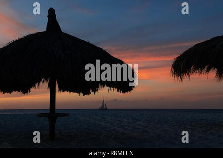 Eagle Beach Aruba au coucher du soleil - un voilier apparaît sous des parasols en chaume avec rose et bleu ciel sur la rive nord-ouest d'Aruba Banque D'Images