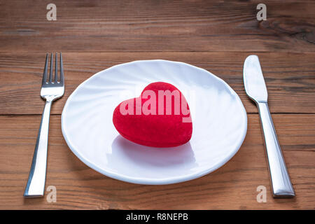 Peluche coeur rouge sur une plaque blanche avec fourchette et couteau sur fond de bois. Symbole de santé minimaliste. La Saint Valentin, Date romantique Table. Banque D'Images