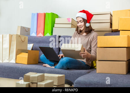 Jeune femme entrepreneur asiatique vendeur en ligne, wearing Christmas hat, travaillant sur son activité en ligne à la maison, vérifier des boîtes de Noël sa prod Banque D'Images