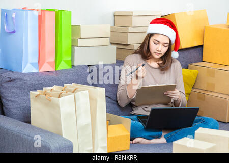 Jeune femme entrepreneur asiatique vendeur en ligne, wearing Christmas hat, travaillant sur son activité en ligne à la maison, vérifier des boîtes de Noël sa prod Banque D'Images