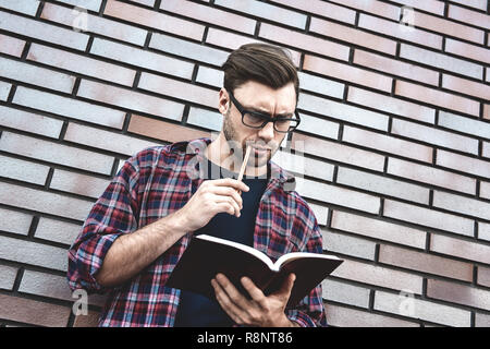 Jeune hipster guy portant des lunettes pour lire un livre ou carnet de notes quelques notes et idées sur mur de brique arrière-plan. Banque D'Images