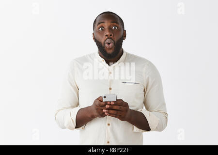 Impressionnés et ravis africains-américains homme barbu adultes lèvres pliage dans l'étonnement et la surprise holding smartphone réagir à awesome news lire en ligne, popping yeux en posant la caméra sur un mur gris Banque D'Images