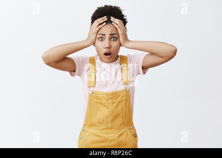 Oh non, je n'ai pas préparé. Choqué et nerveux à la peau sombre mignon étudiante en salopette jaune, tenant les mains sur la tête et haletant avec peur face à l'appareil photo, d'être en situation désespérée Banque D'Images