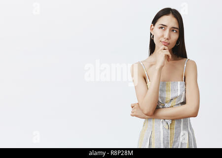 Femme asiatique attrayant créatif designer de vêtements dans l'outfit, tenant la main sur le menton, à la recherche de rêve tout en pensant à des idées ou planifier quelque chose, debout sur un mur gris pensif Banque D'Images