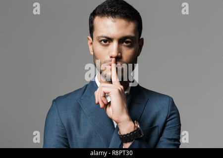 Image de l'homme arabe strict 30s en costume officiel holding index sur lèvres isolated over grey background Banque D'Images