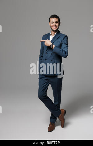 Longueur totale de l'image attractive businessman arabe 30s en costume officiel posant à côté de l'appareil photo avec le doigt sur fond gris isolé Banque D'Images