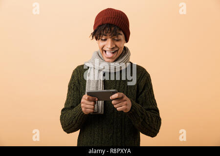 Portrait of african american gamer boy wearing hat and scarf à jouer aux jeux vidéo sur smartphone plus isolé sur fond jaune Banque D'Images