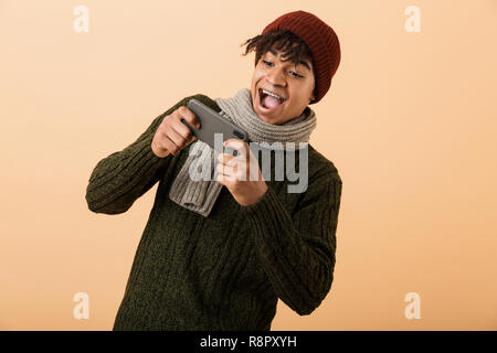 Portrait of african american joyeux gamer boy wearing hat and scarf à jouer aux jeux vidéo sur smartphone plus isolé sur fond jaune Banque D'Images