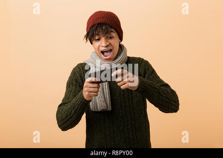 Portrait of african american optimiste gamer boy wearing hat and scarf à jouer aux jeux vidéo sur smartphone plus isolé sur fond jaune Banque D'Images