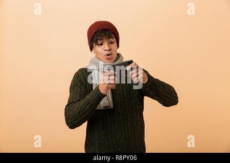 Portrait of african american joyeux gamer boy wearing hat and scarf à jouer aux jeux vidéo sur smartphone plus isolé sur fond jaune Banque D'Images