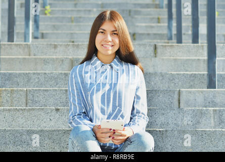 Une jeune fille d'apparence Asiatique, un étudiant vêtue de jeans et une chemise rayée, est titulaire d'une tablette dans ses mains et s'assied sur l'escalier souriant à la ca Banque D'Images