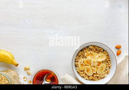 Bol de gruau en céramique blanche avec des tranches de banane et de miel, vu de l'angle élevé en close-up, avec bocal en verre, une cuillère, une banane entière et sur les écrous d'amande Banque D'Images