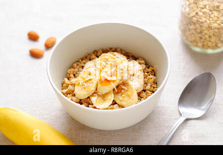 Bol de gruau en céramique blanche avec des tranches de banane, vu de l'angle élevé en close-up, avec bocal en verre, une cuillère, une banane entière et amandes les écrous à l'onglet blanc Banque D'Images