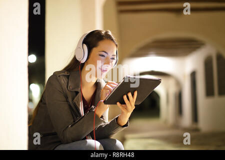 Relaxed woman wearing headphones regarder le support d'un comprimé le soir dans la rue Banque D'Images