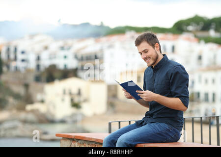 Homme heureux de lire un livre papier assis sur une corniche dans une autre ville en vacances Banque D'Images